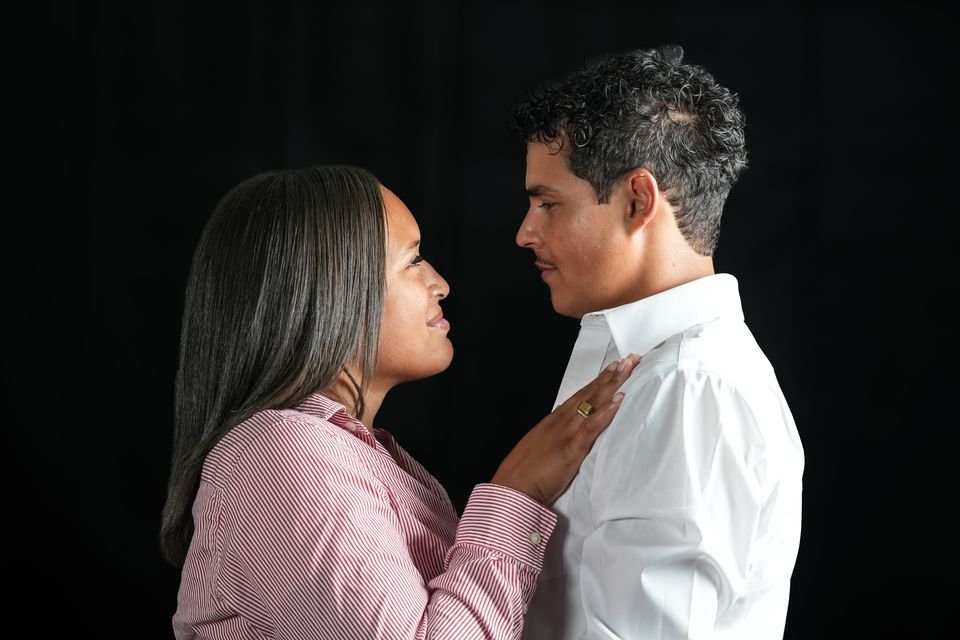 An engaged couple face to face against a black studio backdrop, her hand resting on his chest.