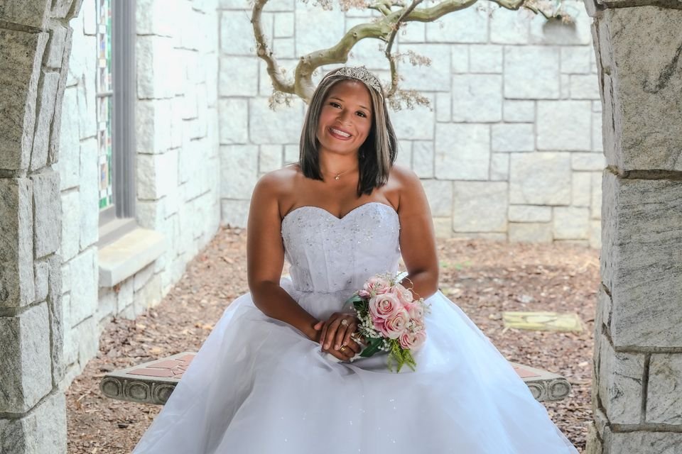 A smiling bride in a strapless white gown and tiara, seated under a stone archway holding a bouquet of pink roses.