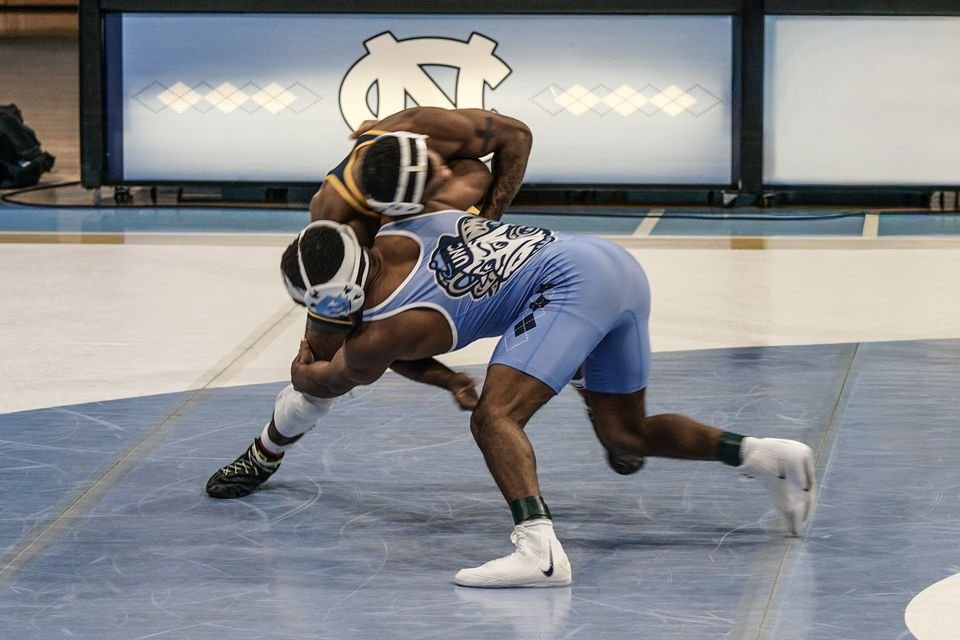 A wrestler in white drives forward while his opponent in maroon fights for position on the mat.
