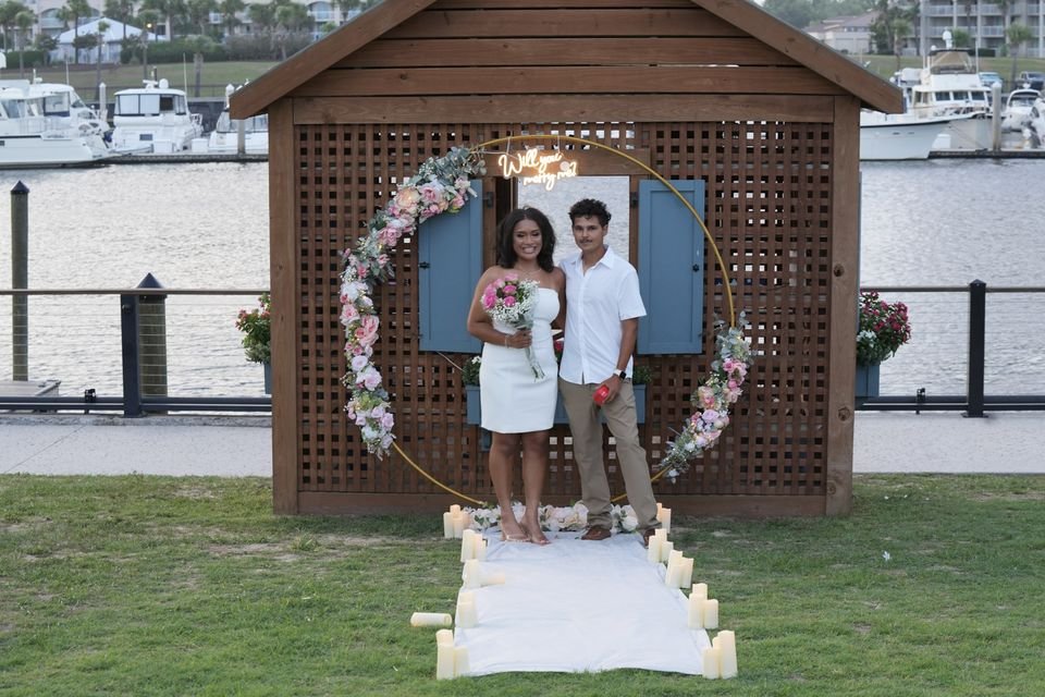 A couple stands under a floral proposal arch on a waterfront lawn with boats in the harbor behind them.